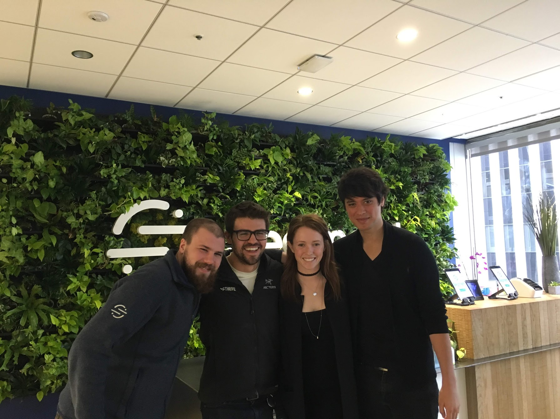 Group of four people posing and smiling in front of a green living wall with a company logo in an office.