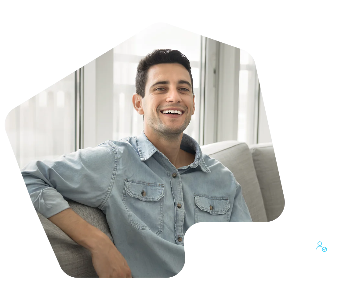 Smiling man in a light blue denim shirt sits on a gray couch in a bright room.