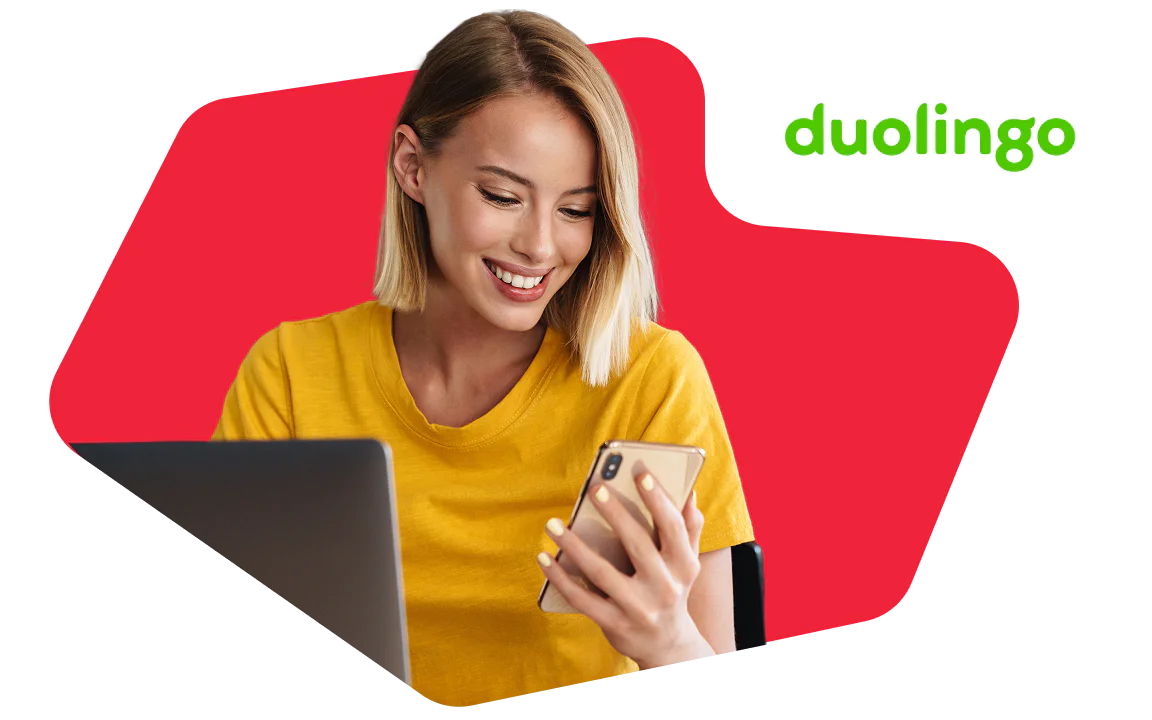 Woman in a yellow shirt smiles at her phone with a laptop open, Duolingo logo in the corner.