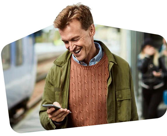 Man in green jacket and orange sweater smiling at his smartphone on a train platform.