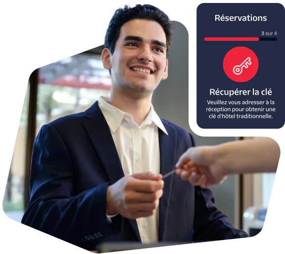 Smiling hotel receptionist gives a room key to a guest with reservation confirmation details on-screen.