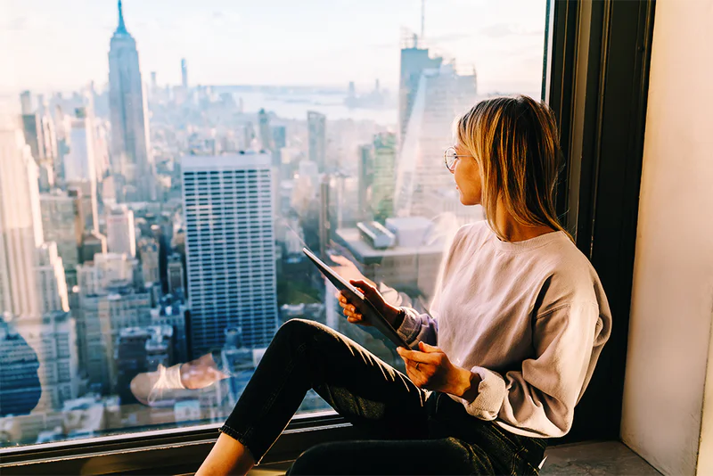 Woman sitting by a window with a tablet, looking out at a city skyline.