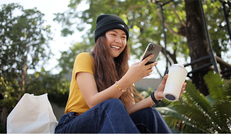 Woman with long brown hair wearing a beanie and yellow shirt, holding a phone and coffee cup, smiling outdoors.