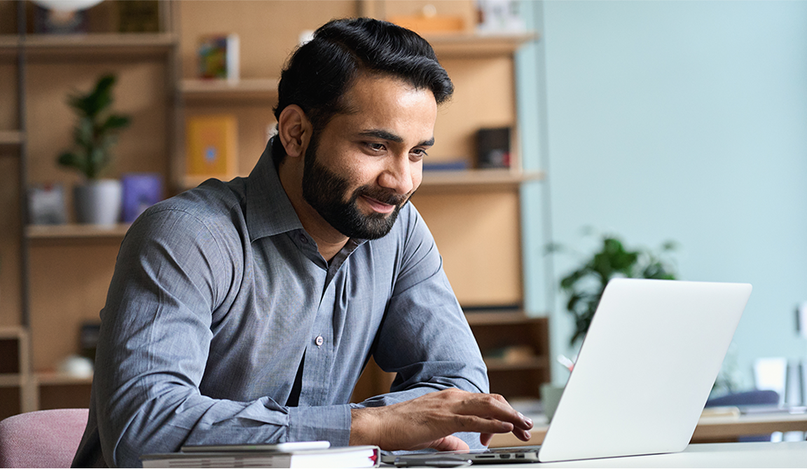 Man in a grey shirt smiling while using a laptop at a desk with books and plants around.