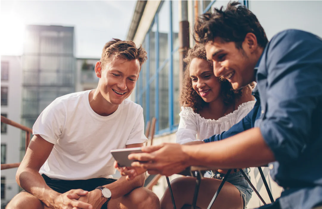 Three friends smiling and looking at a tablet while sitting outside on a sunny day.