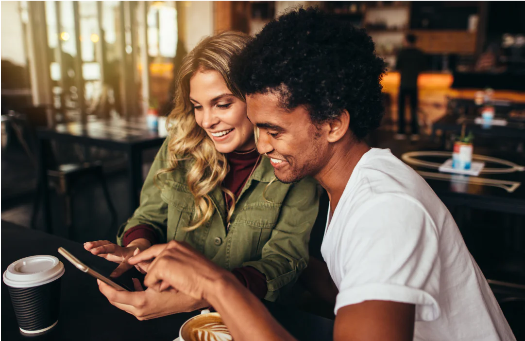 Smiling couple looking at a phone in a café with coffee cups on the table.