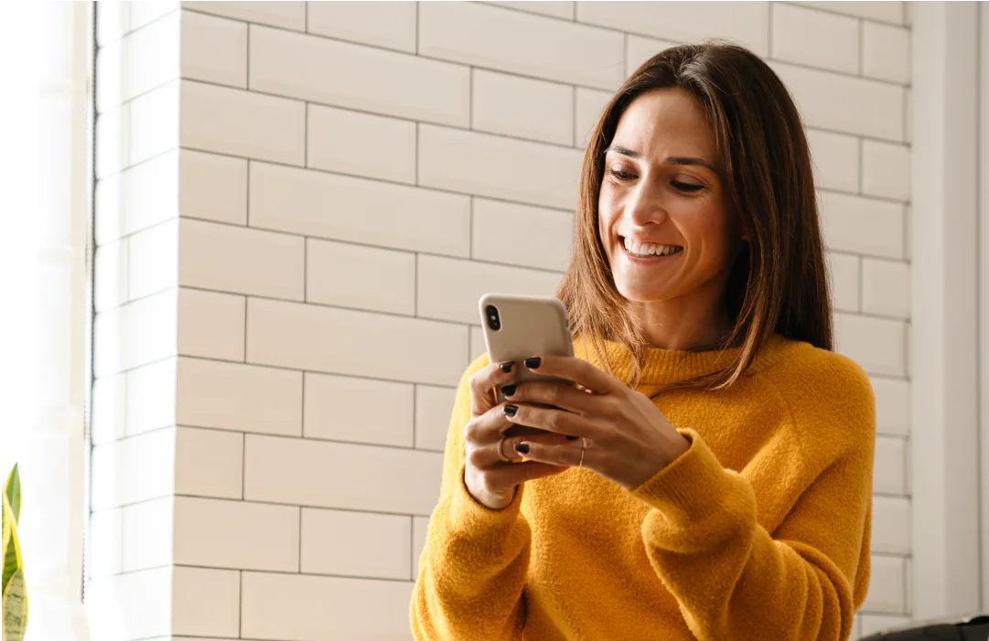Woman in a yellow sweater smiling and looking at her smartphone indoors.