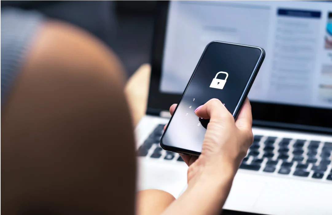 Close-up of a hand entering a passcode on a smartphone with a lock icon, laptop in the background.