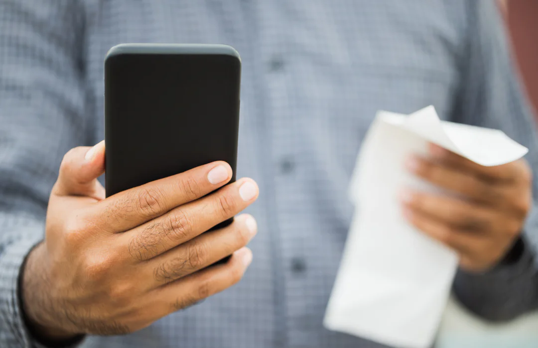 Individual holding a smartphone and a paper receipt with a plaid shirt in the background.