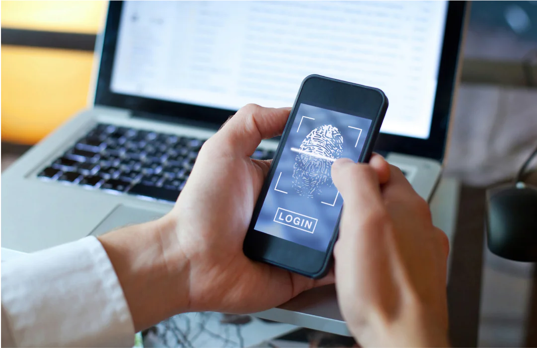 A person using fingerprint scan to log in on a smartphone with a laptop in the background.