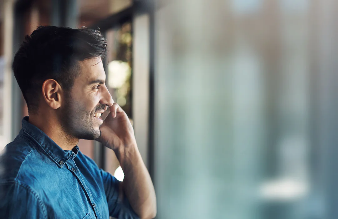 A smiling man holding a phone to his ear while looking out of a window.