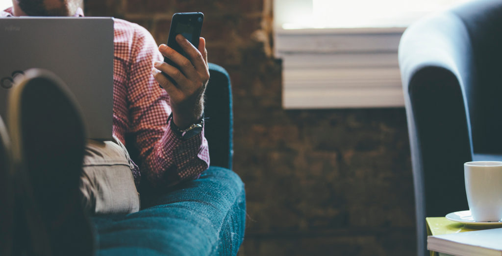 Person sitting on couch with laptop, looking at smartphone in hand, with coffee mug on table.