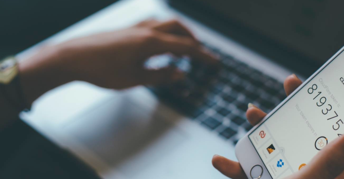 Close-up of hands using a laptop and a smartphone showing a two-factor authentication code