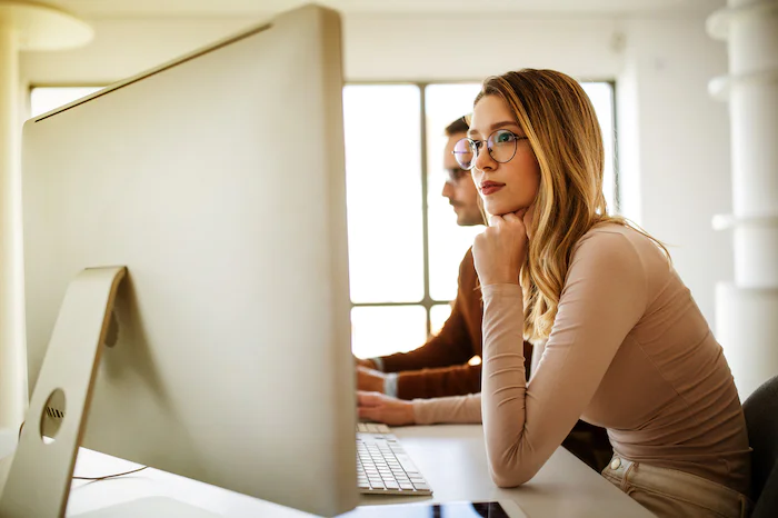 Two professionals working at a desk with large monitors, one wearing glasses, deep in thought.