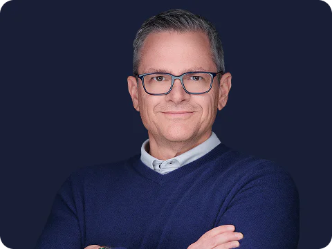 A man with glasses wearing a blue sweater, confidently folding his arms, standing against a dark blue background.
