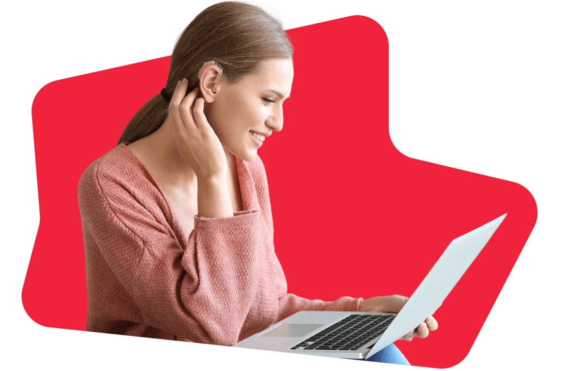 Woman with hearing aid smiling and using a laptop against a red background