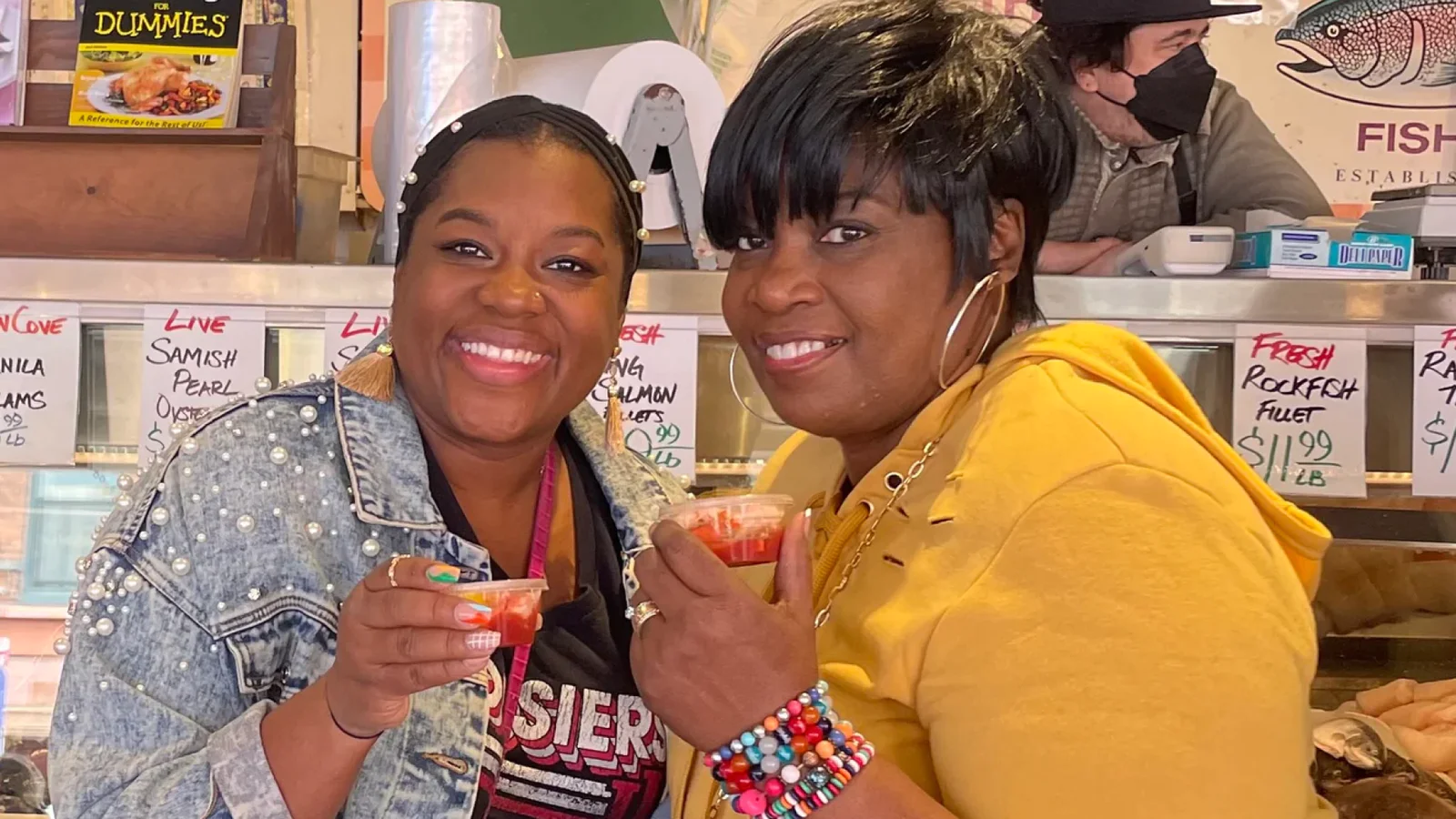 Two women smiling and holding seafood samples in a market.