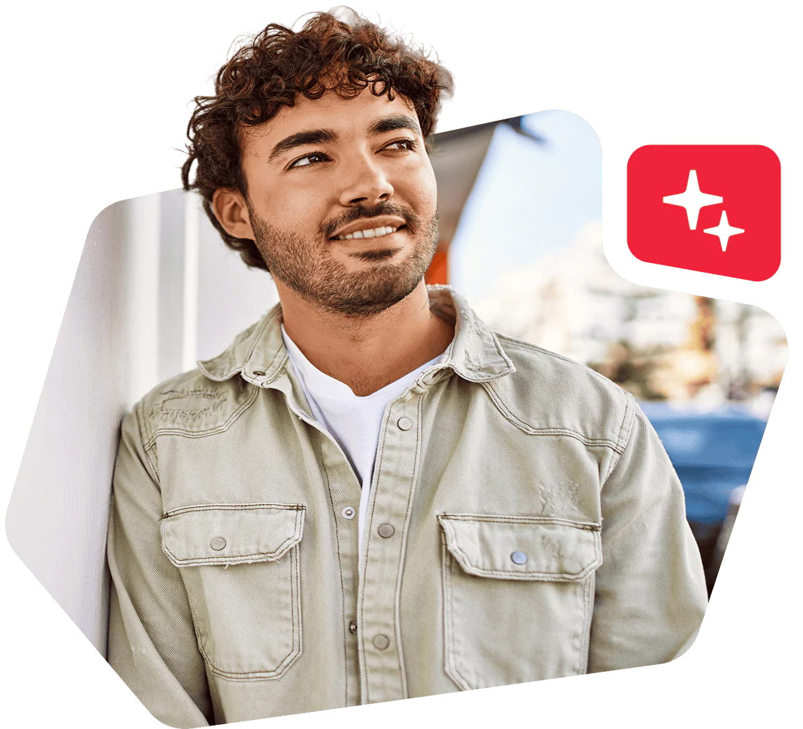 Man with curly hair and a beard smiling while leaning against a white wall, wearing a light green shirt.