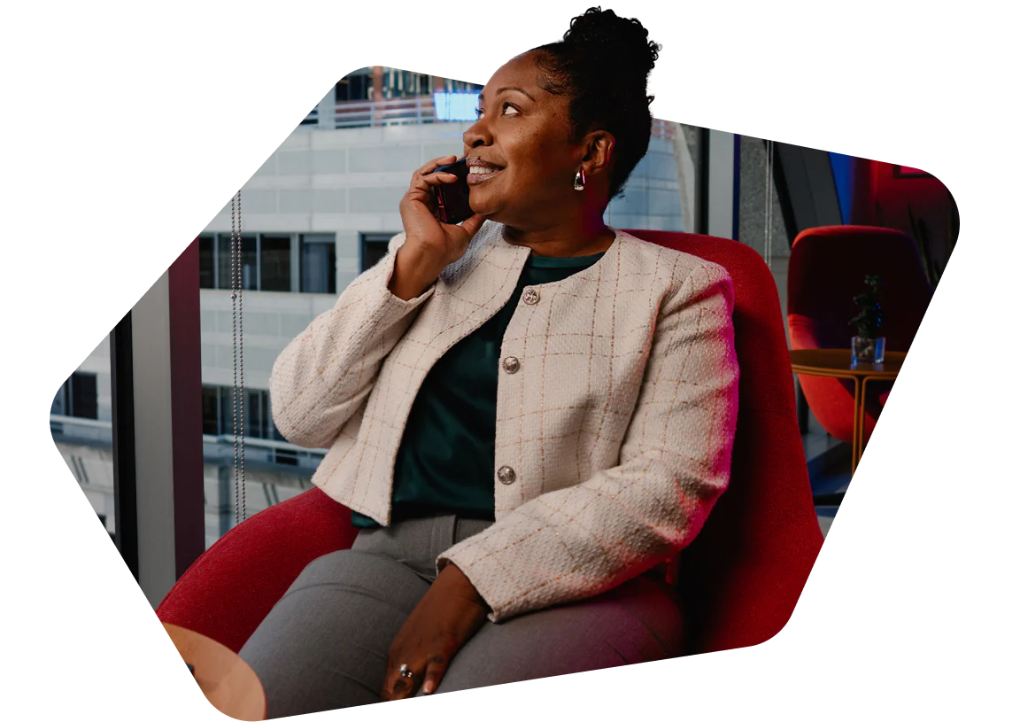 Woman in a white blazer sitting in a red chair, talking on a phone with a cityscape in the background.