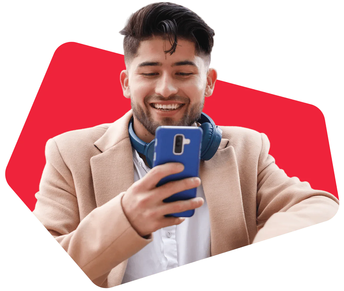 Smiling man with beard using phone, wearing headphones and a beige blazer against red background.