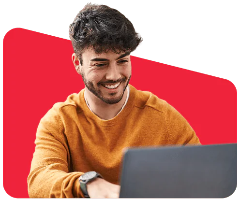 Man in an orange sweater smiling while using a laptop against a red background.