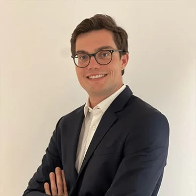 Man wearing glasses and a dark suit with arms crossed, smiling against a plain white background.