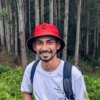 Man in red hat and white shirt smiling in front of a forested area with trees and greenery.