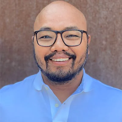 Bald man with glasses and beard smiling, wearing a light blue shirt, against a brown textured background.