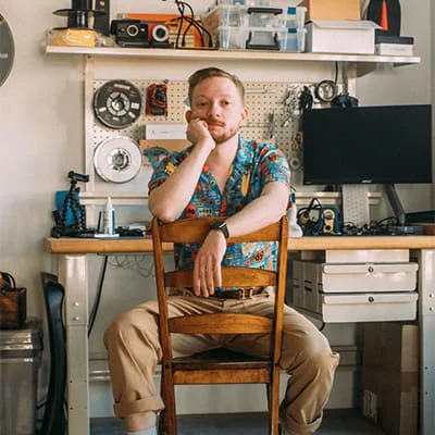 Man in a floral shirt sitting on a wooden chair in a workshop with tools and electronics.