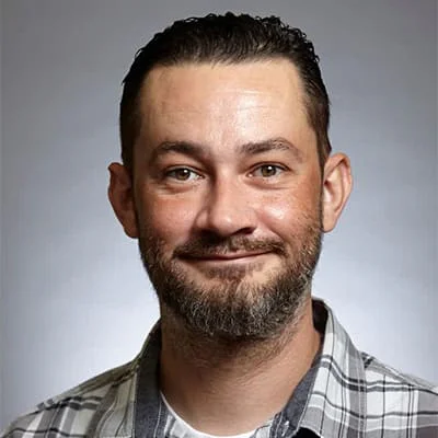 Close-up of a smiling man with a groomed beard wearing a checkered shirt, gray background.