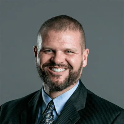 A man with a beard, dressed in a suit and tie, smiling against a gray background.