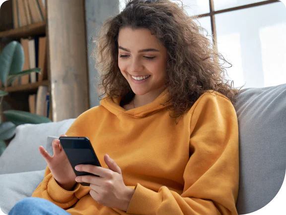 Woman with curly hair in a yellow hoodie smiling while using a smartphone on a sofa.
