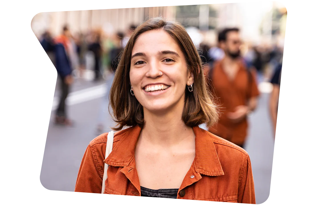 Smiling Woman in Burnt Orange Jacket at Outdoor Event Woman with brown hair and a nose ring smiles while standing outside at a busy event.
