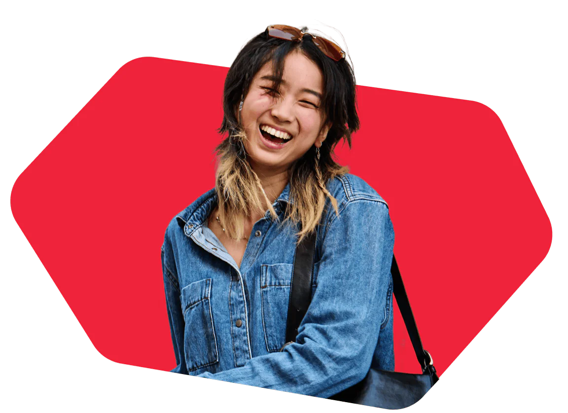 Woman with sunglasses on her head, wearing a denim jacket, smiling in front of a red background.