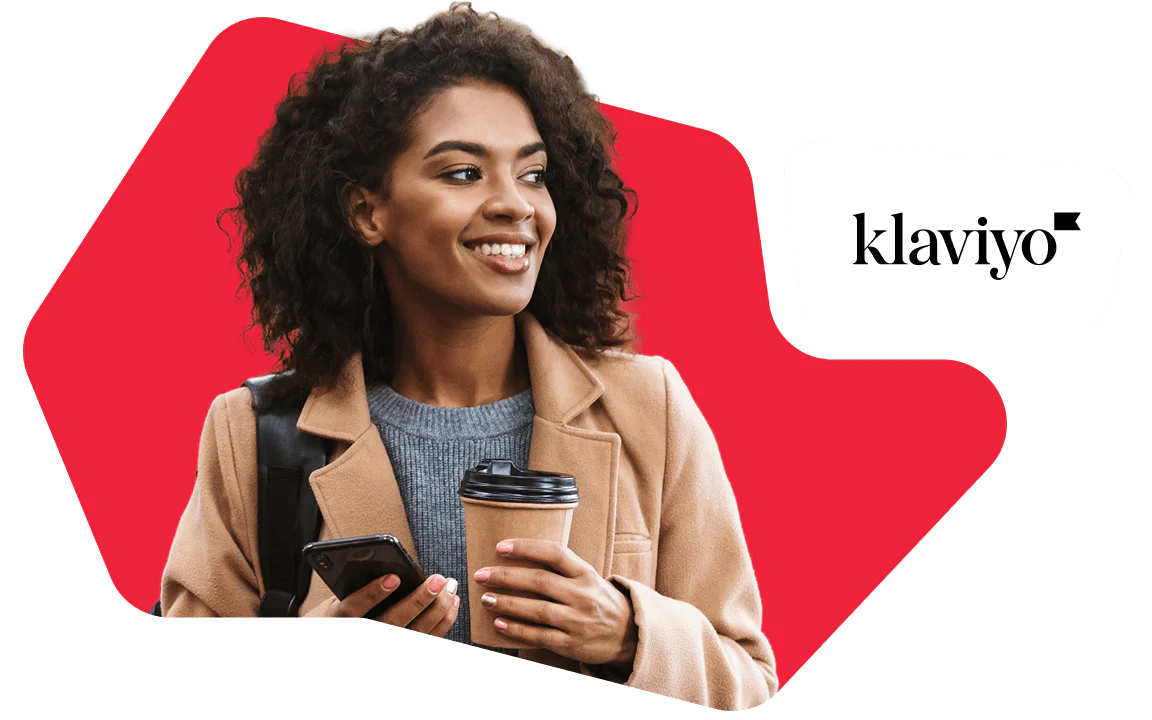 Woman Holding Coffee and Phone with Klaviyo Logo Woman with curly hair holding a coffee cup and smartphone, wearing a tan coat, smiling.