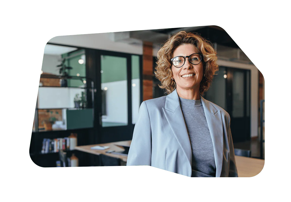 Smiling woman with curly hair and glasses in a blue blazer, standing in a modern office.