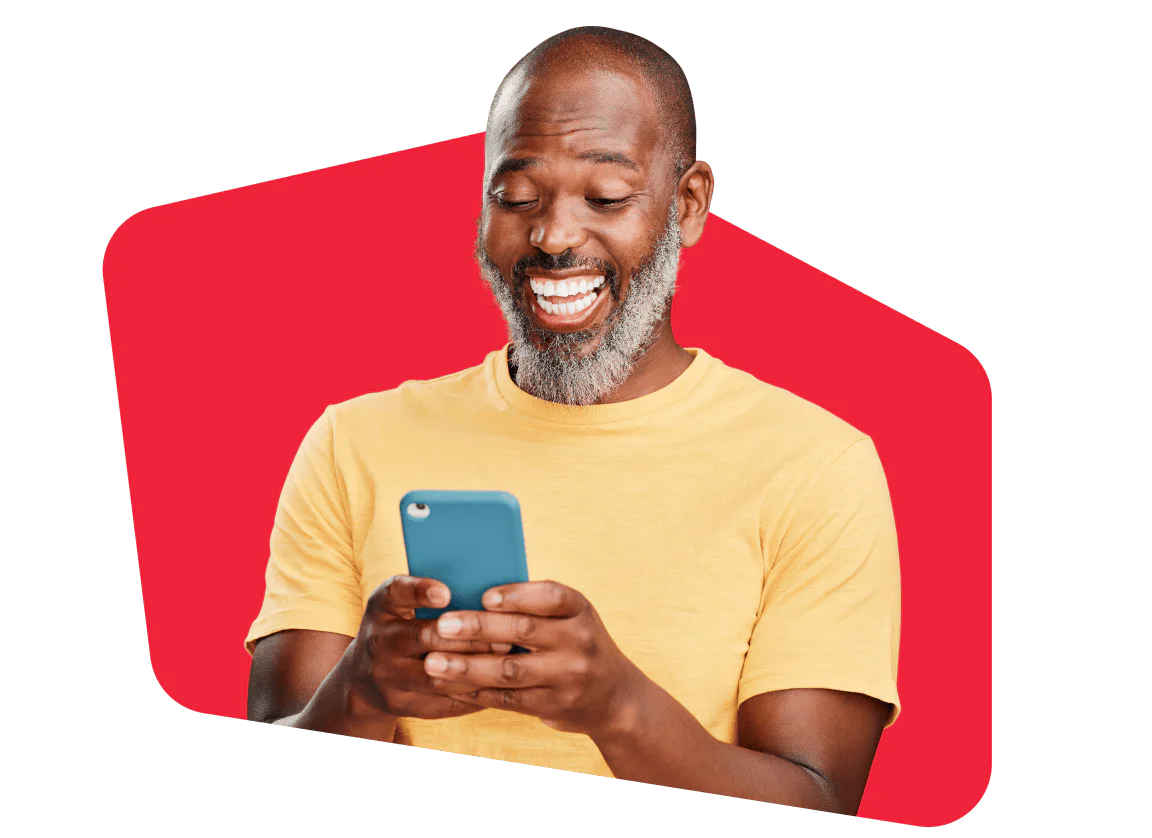 Smiling man with a gray beard, wearing a yellow shirt, holding and looking at a smartphone, red background.