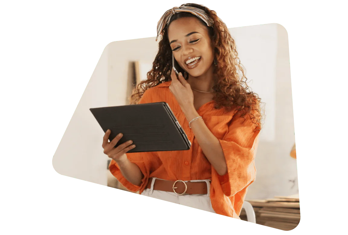 Smiling woman in an orange shirt using a tablet and talking on the phone.