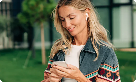 Trust the process: How branded communications drive consumer confidence Woman with earbuds smiling while looking at her smartphone outside in a green setting.