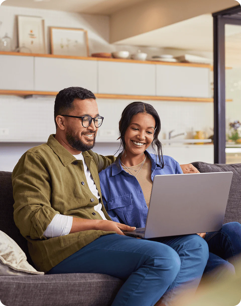 A couple sits on a couch smiling while looking at a laptop in a modern living room setting.