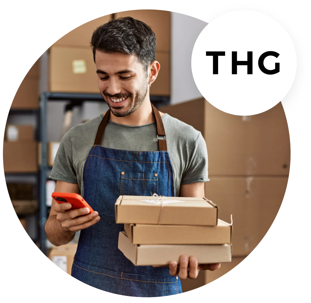 Man in apron holding packages and looking at phone, standing in a warehouse, THG logo in the corner