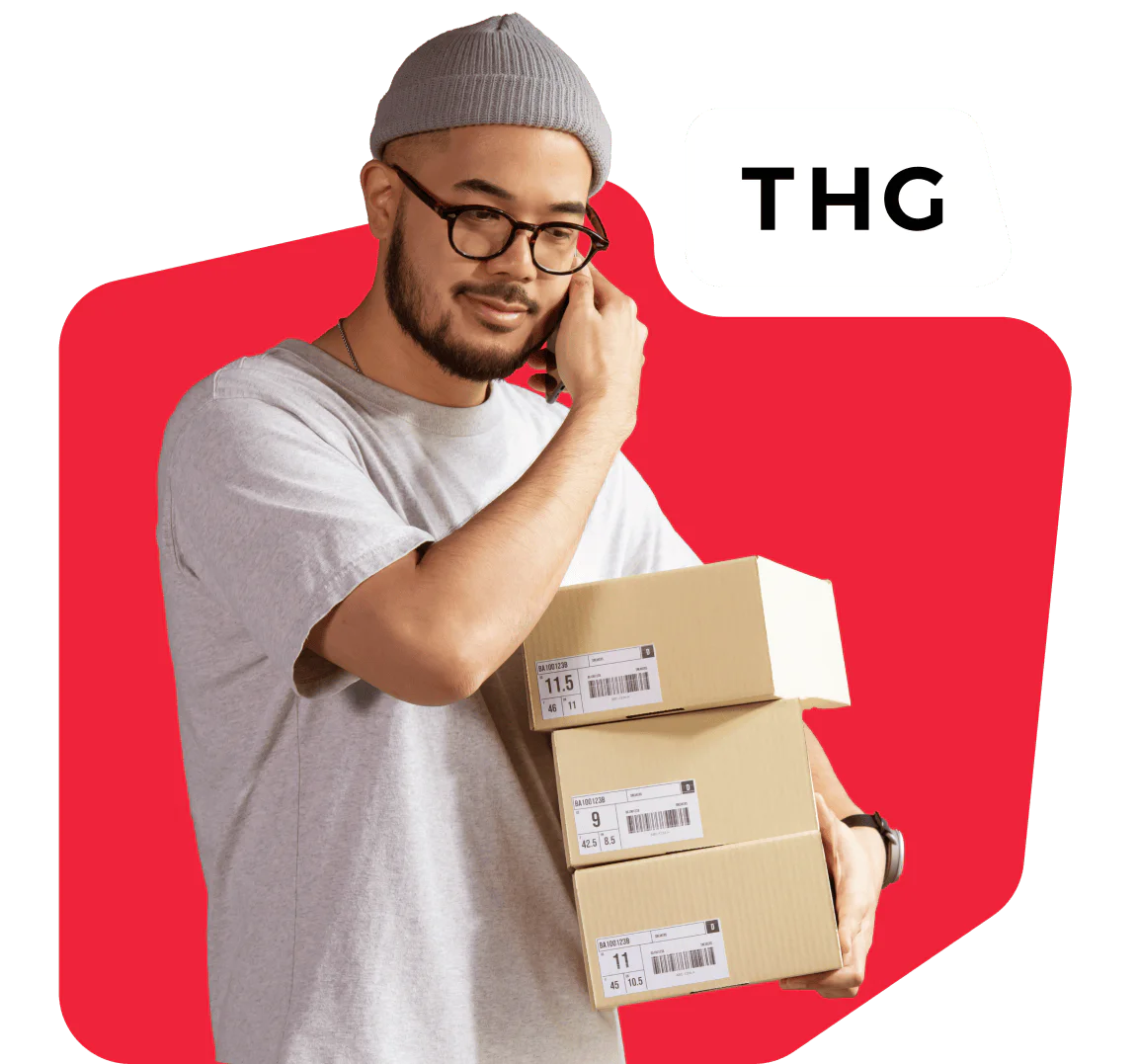 Man Holding Boxes with THG Logo in Background Man in grey beanie and glasses holding packages with THG logo on red background.