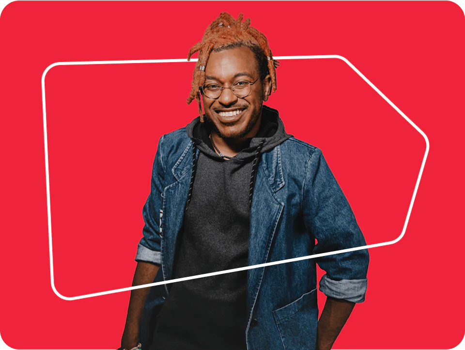A man with dreadlocks, glasses, and a denim jacket over a black hoodie smiles against a red background.