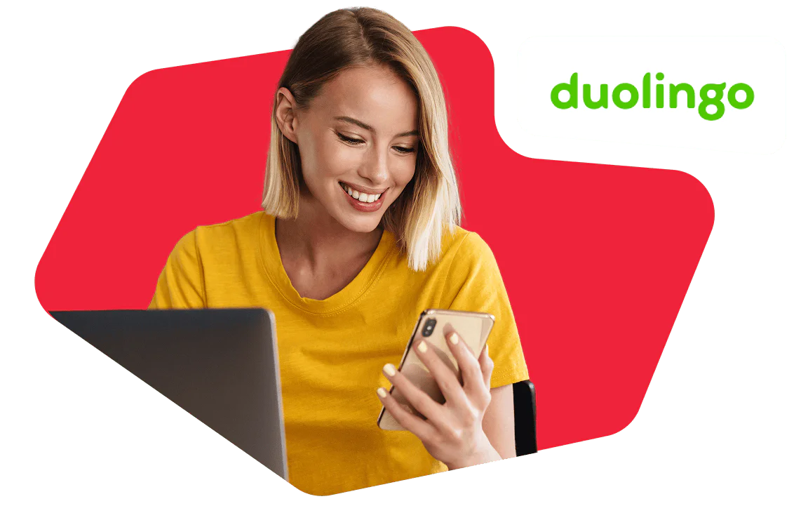 Woman in a yellow shirt smiles at her phone with a laptop open, Duolingo logo in the corner.