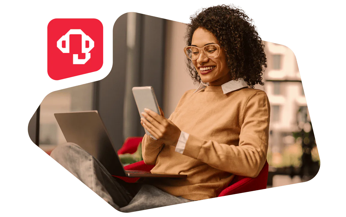 Woman with curly hair in glasses using a smartphone and laptop, smiling in a cozy indoor setting.