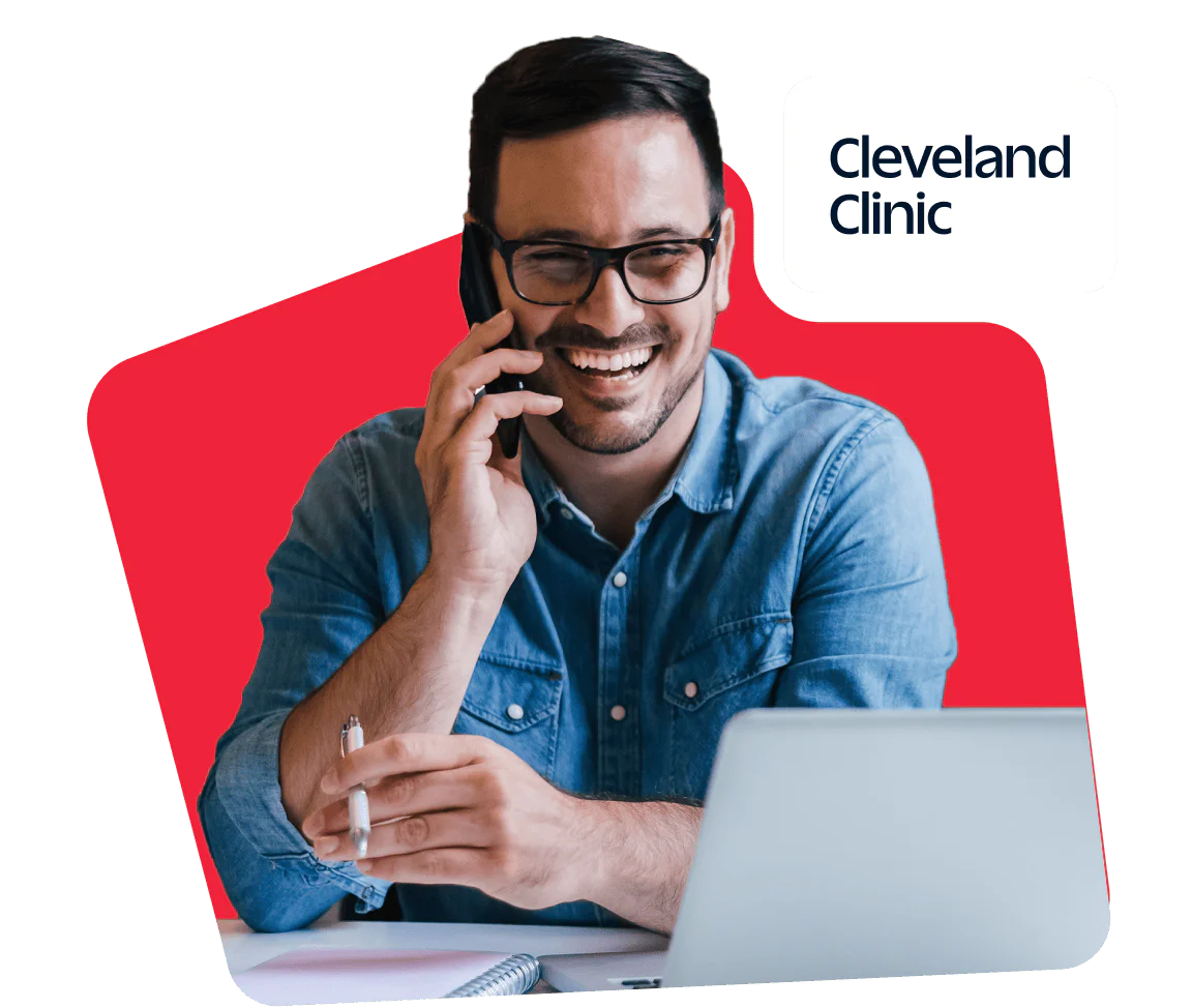 Smiling man talking on phone while using laptop with Cleveland Clinic logo in background.
