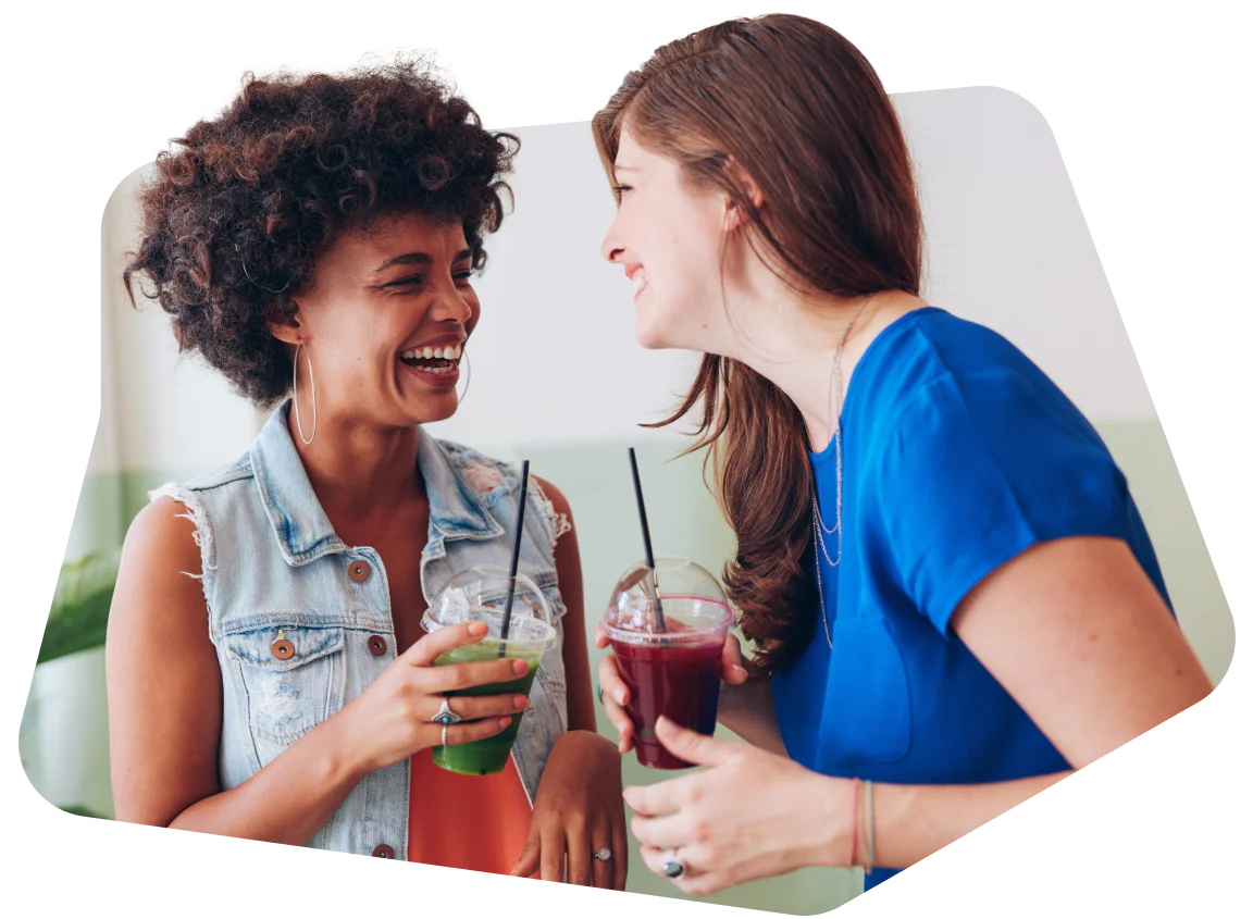 Two women smiling and holding drinks while talking inside a cafe.